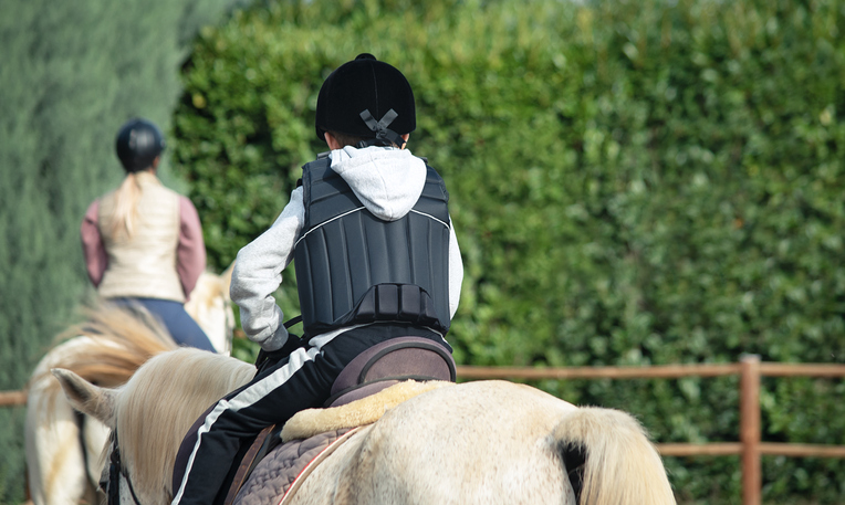 Back view of kid riding white horse during horseback lessons.