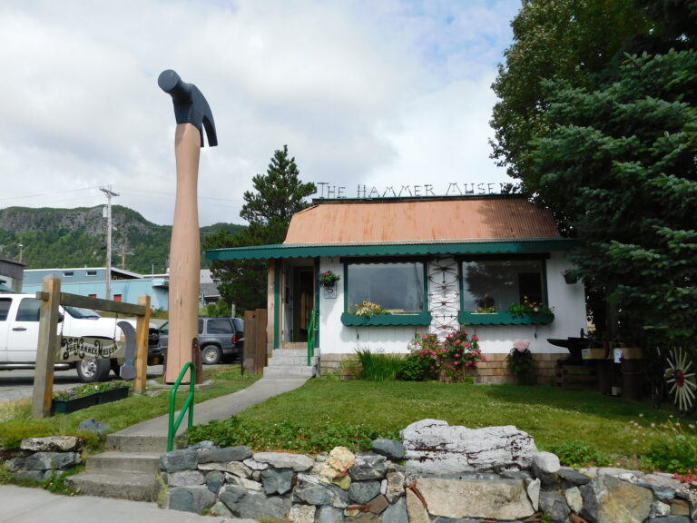 An exterior view of the hammer museum, a small white building with green trim. In front, a huge hammer stands tall on the lawn. In the back, Alaska's rolling hills.