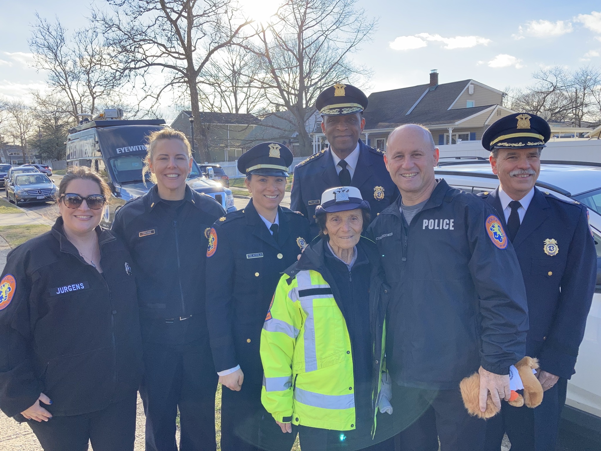 91-year-old crossing guard Louise Kobs wears a bright yellow-green jacket and stands with a group of smiling police officers